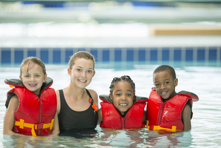 Life gaurd instructor in a swimming pool with 3 children smiling. The 3 children has on life jackets.